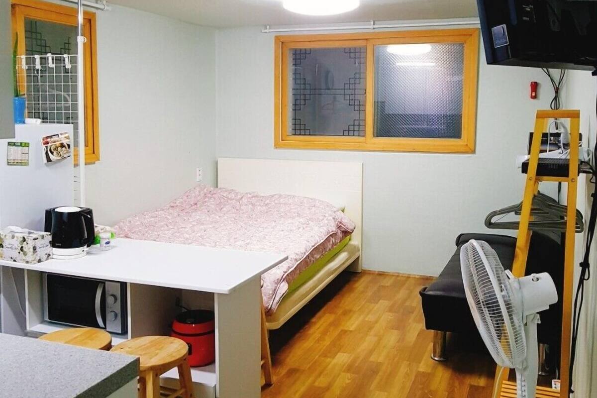 A compact dormitory room with a bed, a desk, a chair, a TV, and a fan.
