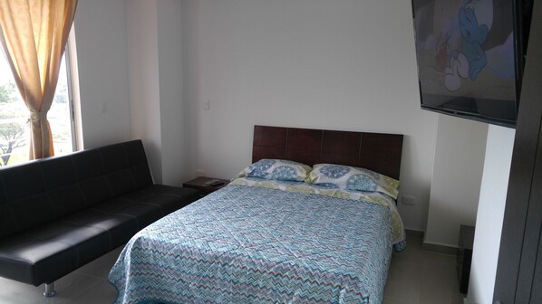 A bedroom with a bed, a black sofa, a TV on the wall, and a window with curtains.