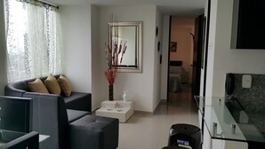 Un salon moderne avec un canapé, une table basse et un vase décoratif. Un miroir est accroché au mur et on aperçoit un coin cuisine à l’arrière-plan.