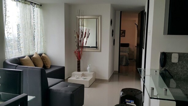 Ein modernes Wohnzimmer mit einer Couch, einem Couchtisch und einer dekorativen Vase. An der Wand hängt ein Spiegel, im Hintergrund ist eine Küche zu sehen.