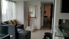 A modern living room with a sofa, coffee table, and a decorative vase. There is a mirror on the wall and a kitchen area visible in the background.