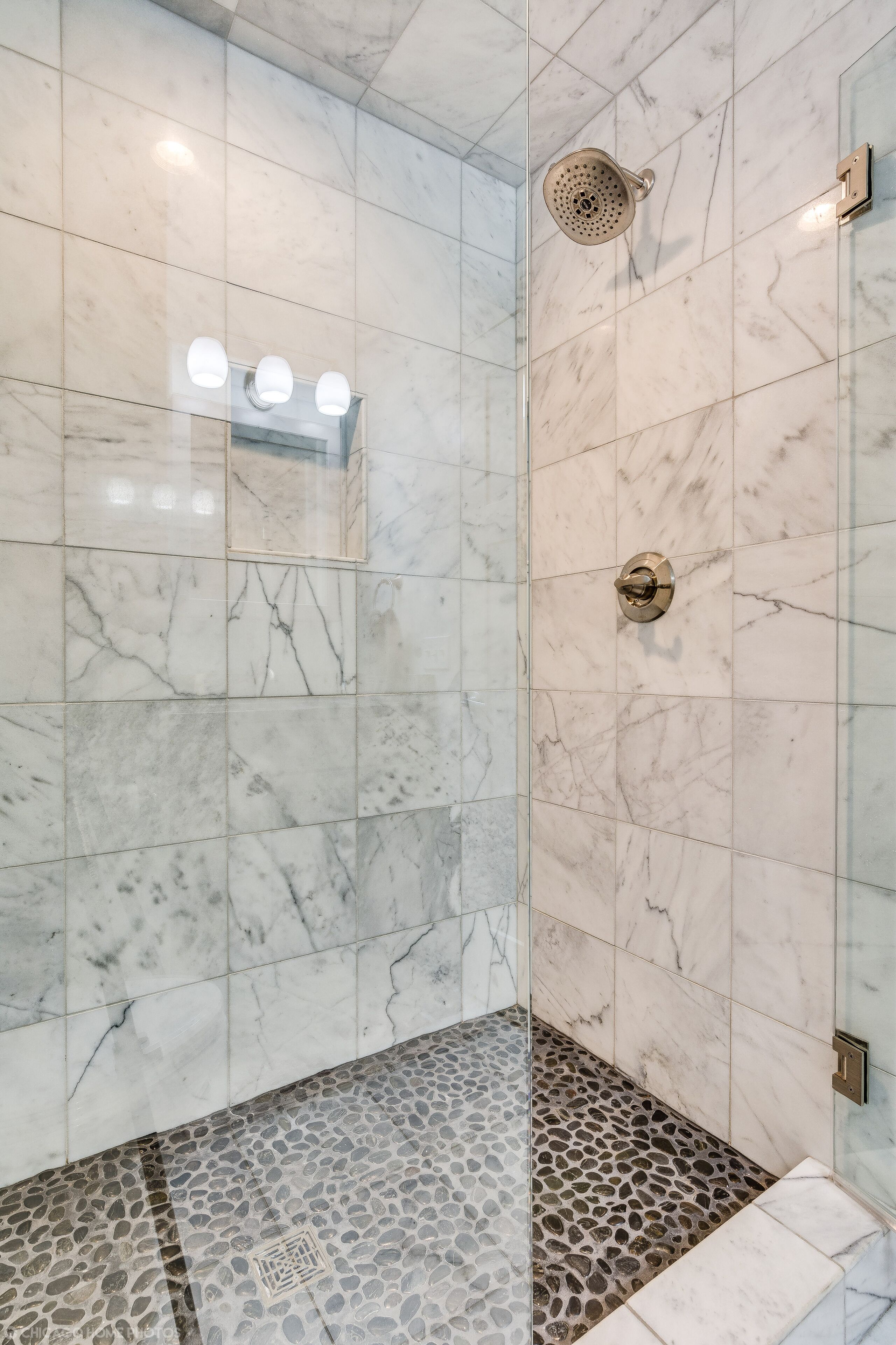 Un baño moderno con paredes de mármol, ducha con cabezal de lluvia y piso con diseño de guijarros.
