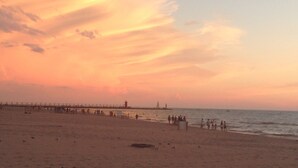 Ein Strand mit einem Steg, der sich ins Wasser erstreckt, Menschen, die am Ufer entlanggehen, und einem Sonnenuntergang im Hintergrund.