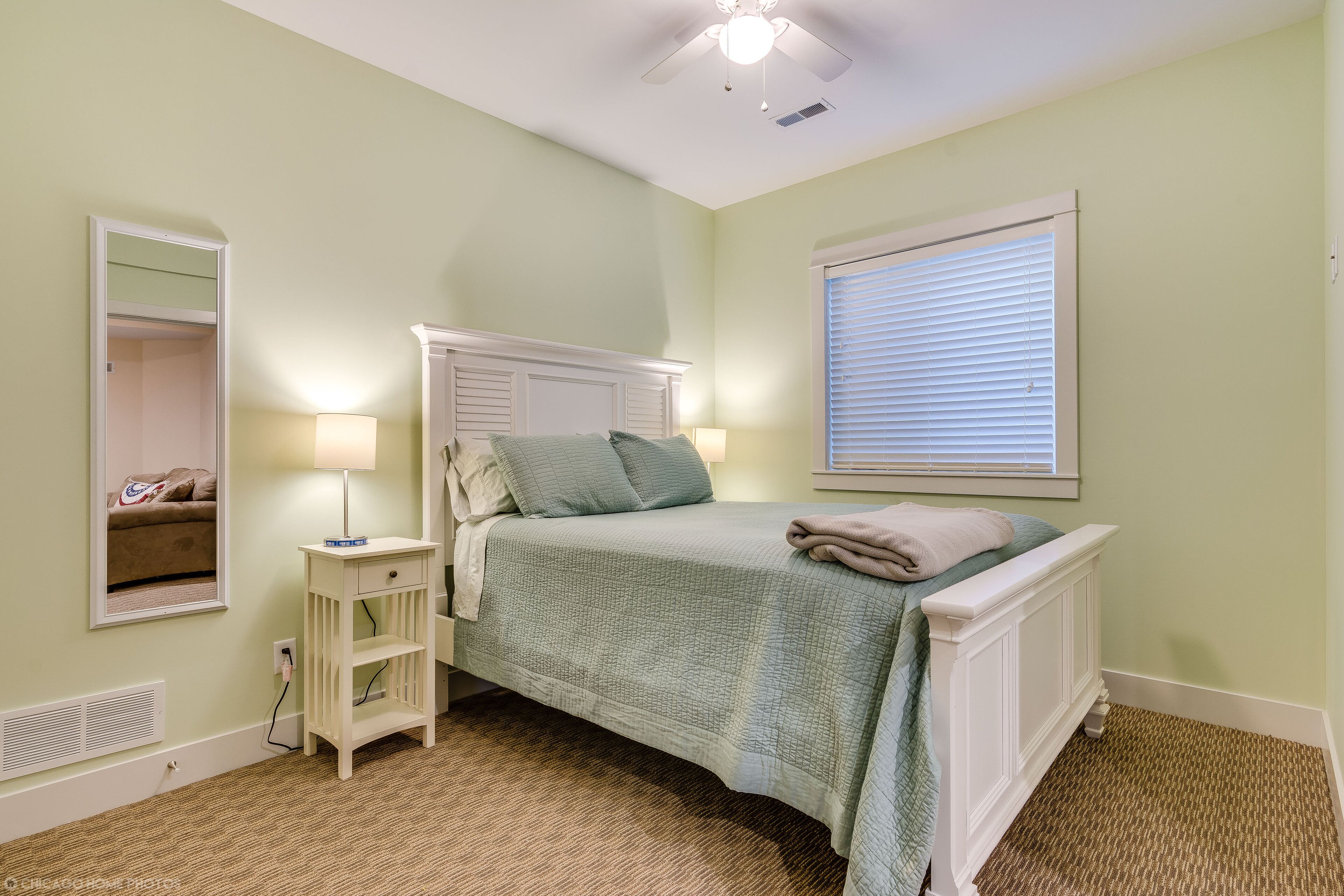 A bedroom with a bed, bedside lamps, a nightstand, a window with blinds, and a ceiling fan.