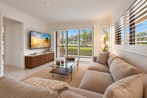 Una sala de estar moderna con un televisor de pantalla plana grande, una mesa de centro de vidrio, un sofá color beige y vistas amplias por ventanas grandes.