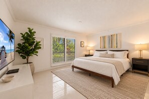 Un dormitorio con cama, mesitas de noche, televisor, planta y ventana con persianas.