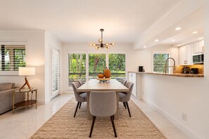 A modern dining area with a large table, chairs, a chandelier, and a view through large windows.
