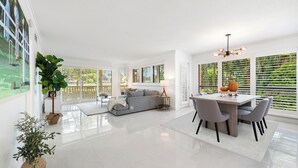A spacious living area with a grey sofa, a dining table with chairs, and a large window with blinds.