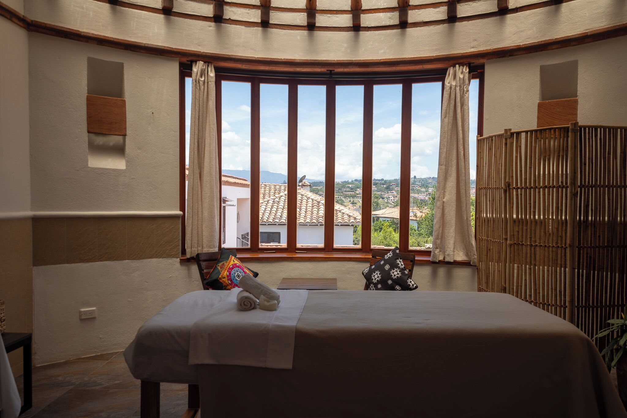 A bedroom with a bed, a window with curtains, a view of buildings, and a bamboo screen.