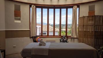 A bedroom with a bed, a window with curtains, a view of buildings, and a bamboo screen.