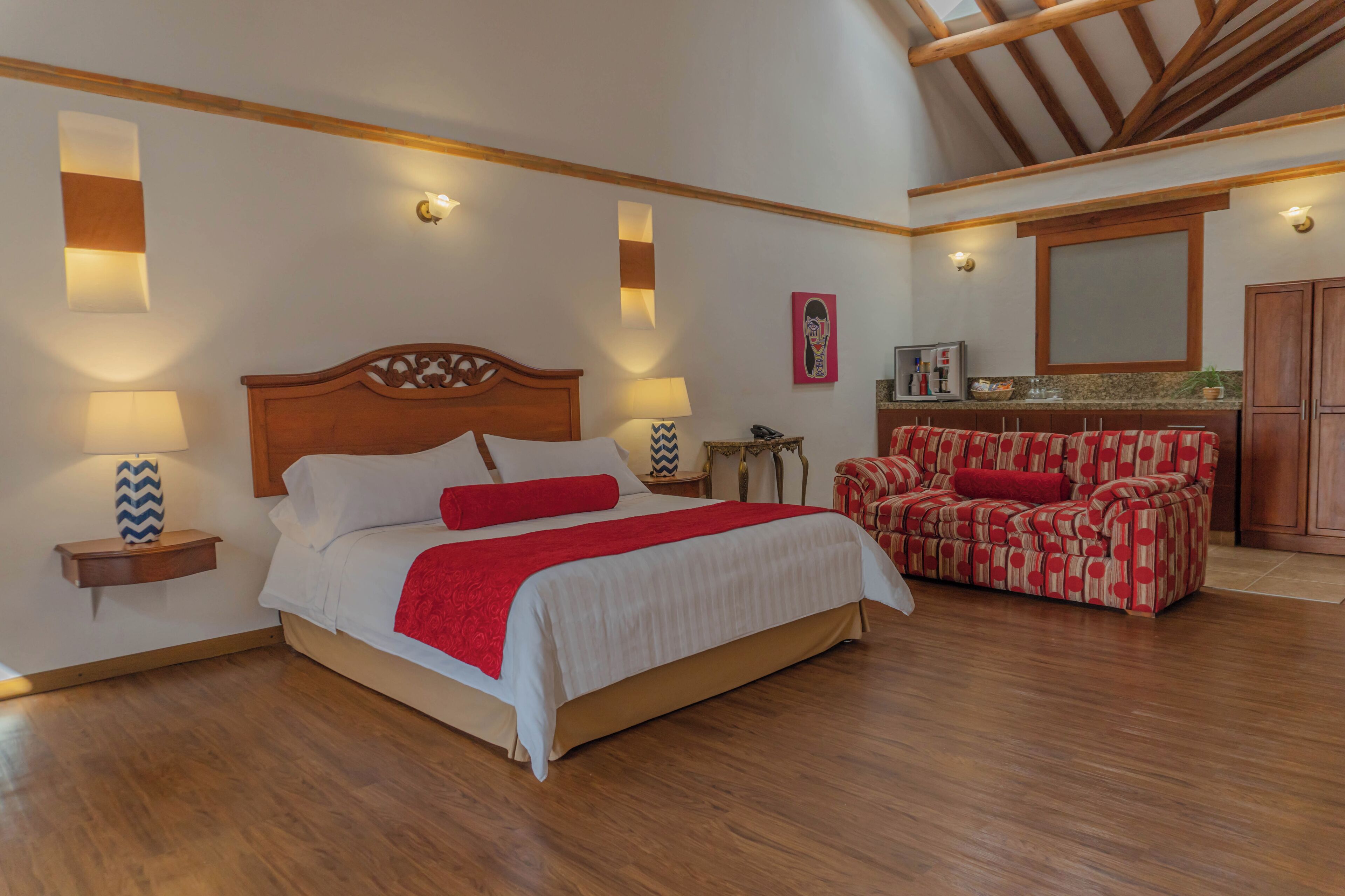 Un dormitorio con una cama de madera, lámparas de noche, un sofá rojo y una cocina al fondo.