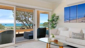 Sala de estar moderna com mesa de centro de vidro, sofá com almofada de 'welcome' e uma grande janela com vista para a praia e o oceano.
