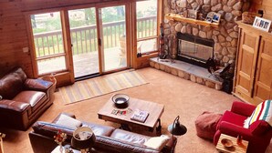 Un acogedor interior de cabaña con chimenea de piedra, mobiliario de cuero y grandes ventanas que ofrecen vistas del exterior.