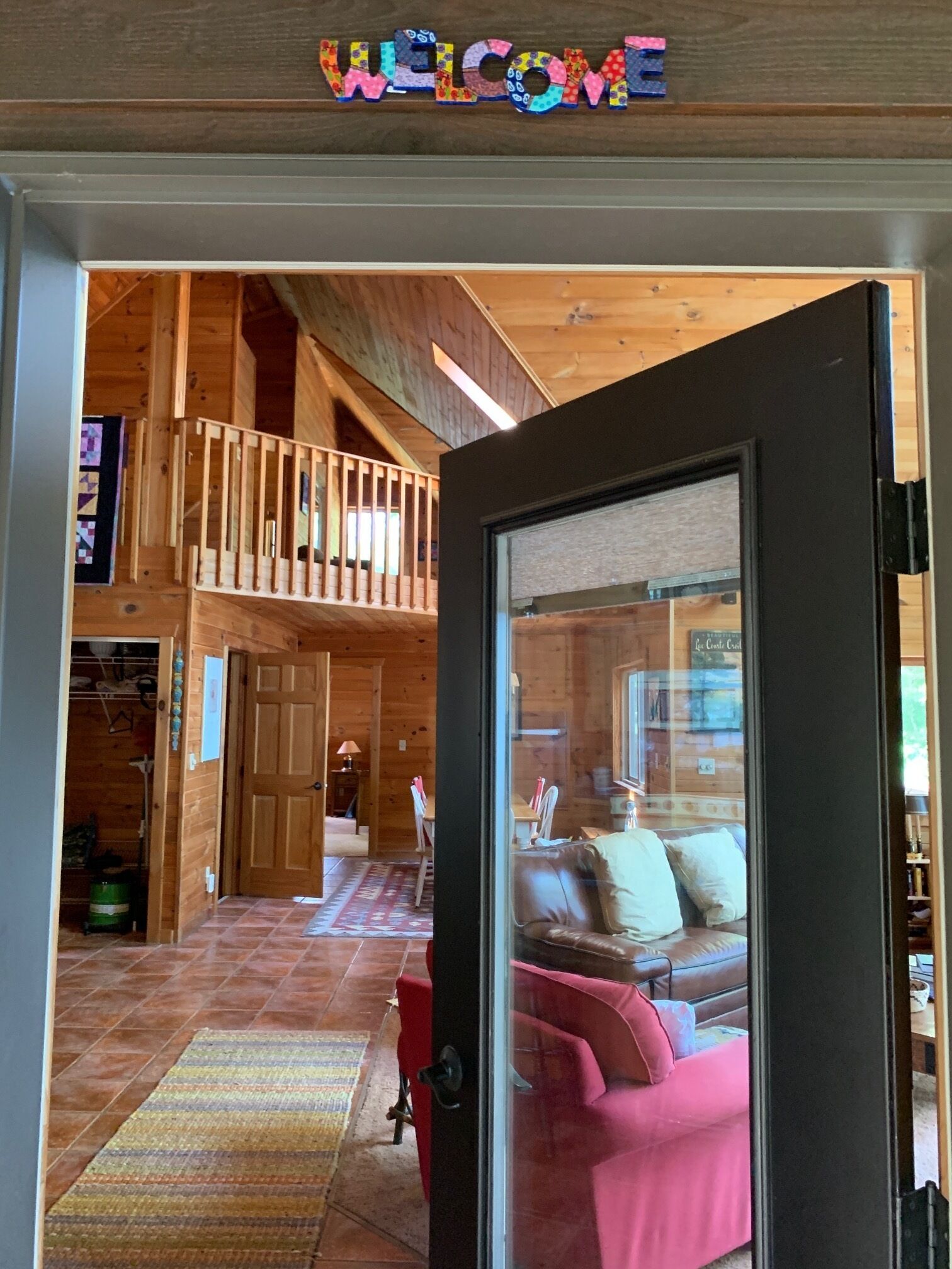 A wooden interior with a 'WELCOME' sign, a staircase, a dining area, and a living room with a pink sofa.