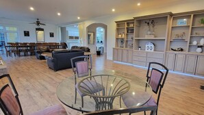 Un vaste salon comprenant une table à manger avec un plateau en verre, un canapé et une cheminée. Il y a également un coin cuisine avec des meubles et des étagères en bois.