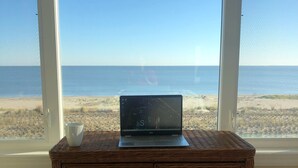 Um laptop sobre uma escrivaninha de vime, com vista para a praia e o oceano através de janelas amplas.