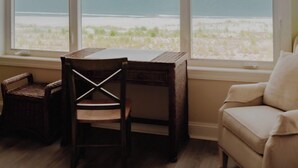 Quarto com uma grande janela com vista para a praia, uma mesa de madeira, uma cadeira de vime e um sofá bege.