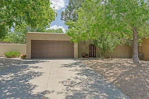 A residential garage with a brown door, a small garden area with a potted plant, and a tree providing shade.