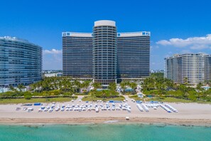 Ein Hotel direkt am Strand mit einer geschwungenen Fassade, mehreren Balkonen und einem Poolbereich mit Sonnenschirmen.