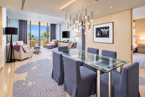 Habitación de hotel moderna con una mesa de comedor de vidrio, asientos cómodos, un televisor de pantalla plana y un ventanal con vistas a palmeras.