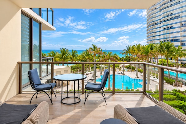 A balcony with a view of a pool and palm trees, a round table, and two chairs.