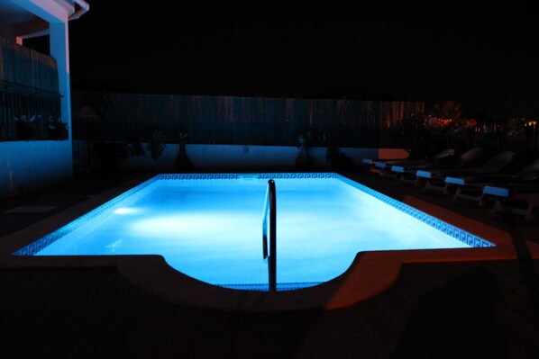 A swimming pool at night with a clear water surface, surrounded by lounge chairs and a low wall.