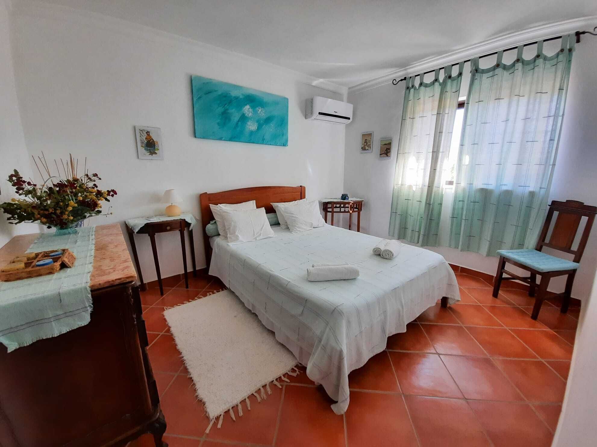 Ein ordentlich eingerichtetes Schlafzimmer mit einem Bett, einem kleinen Tisch mit Blumenvase, einem Sessel und einem Fenster mit Vorhängen.