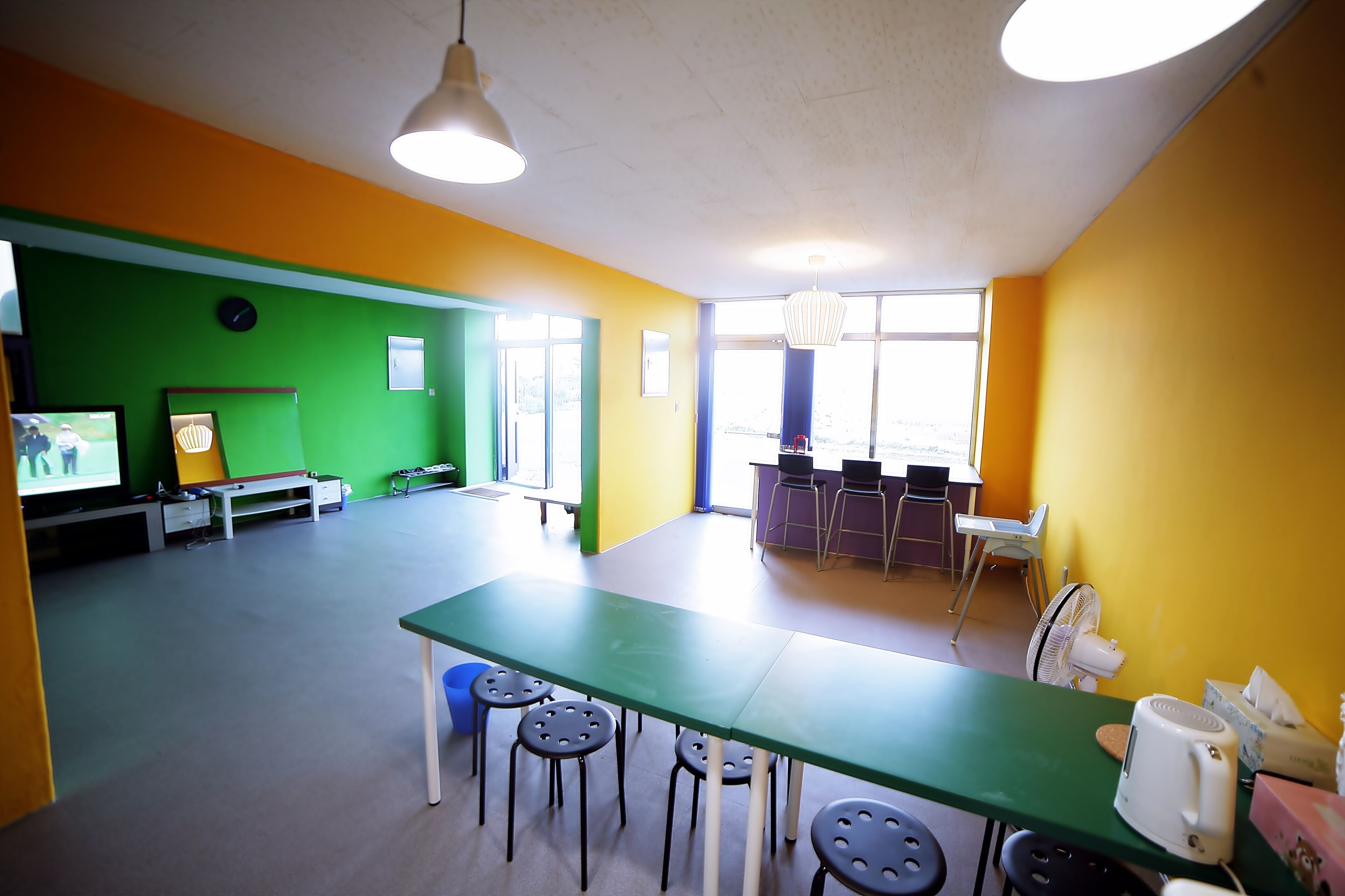A brightly colored room with a green table and black stools, a television, and a fan.