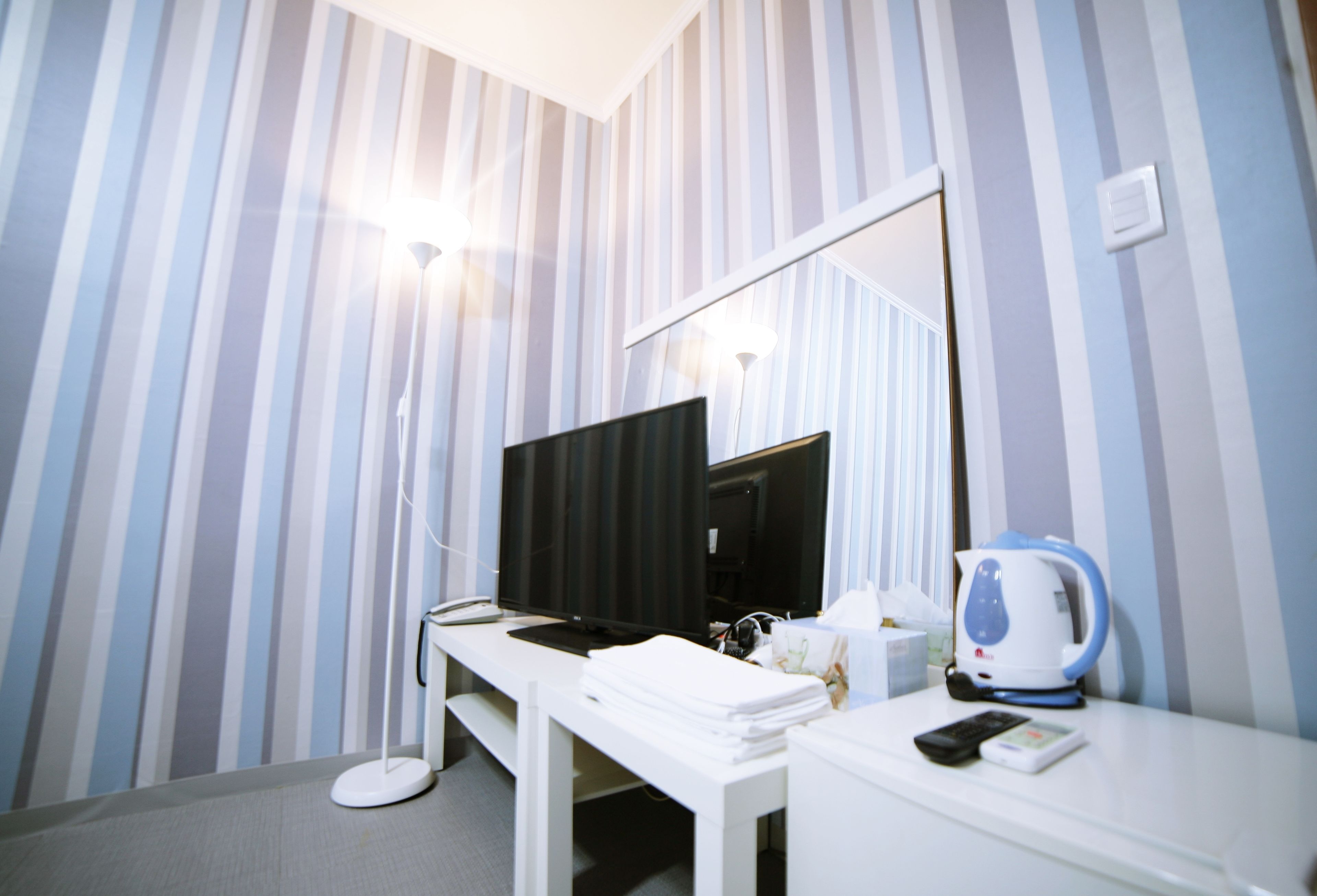 Une chambre d’hôtel avec un mur à rayures, un bureau avec un ordinateur, une bouilloire et une table de chevet avec un téléphone et des mouchoirs.