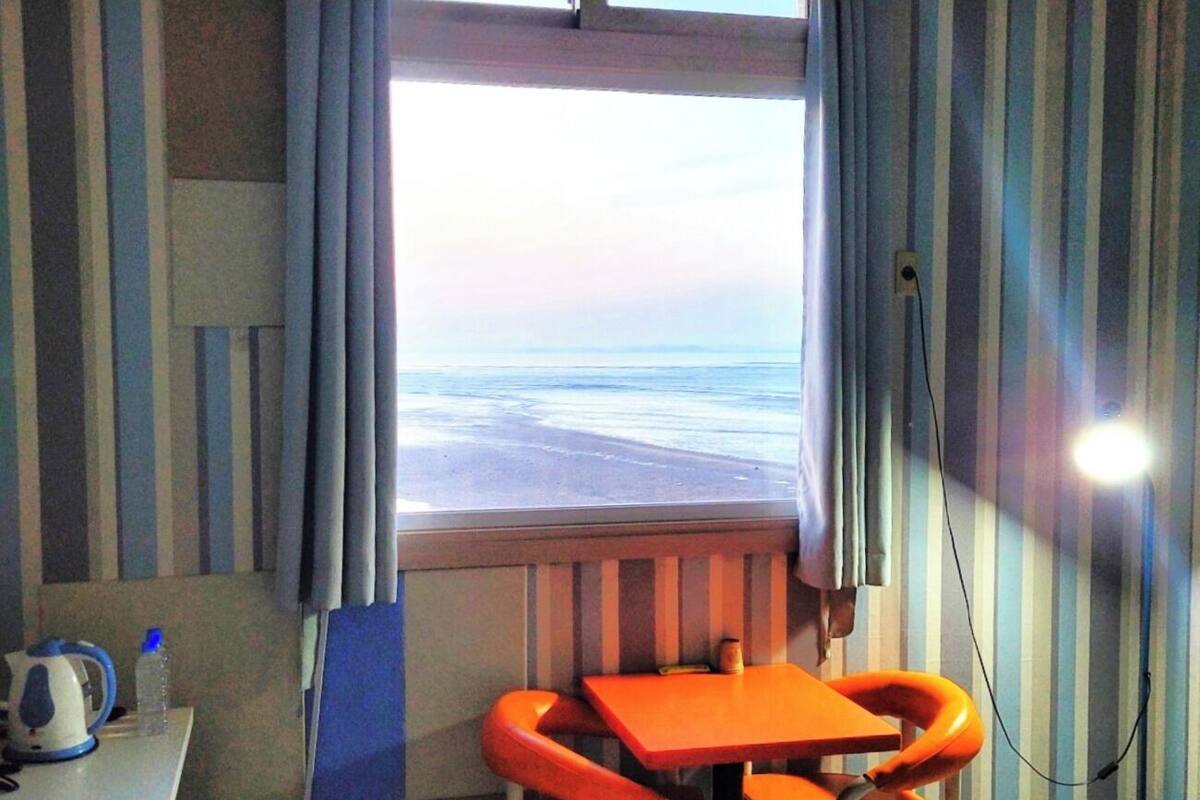 A room with a window overlooking a beach, a table with a kettle, and striped curtains.