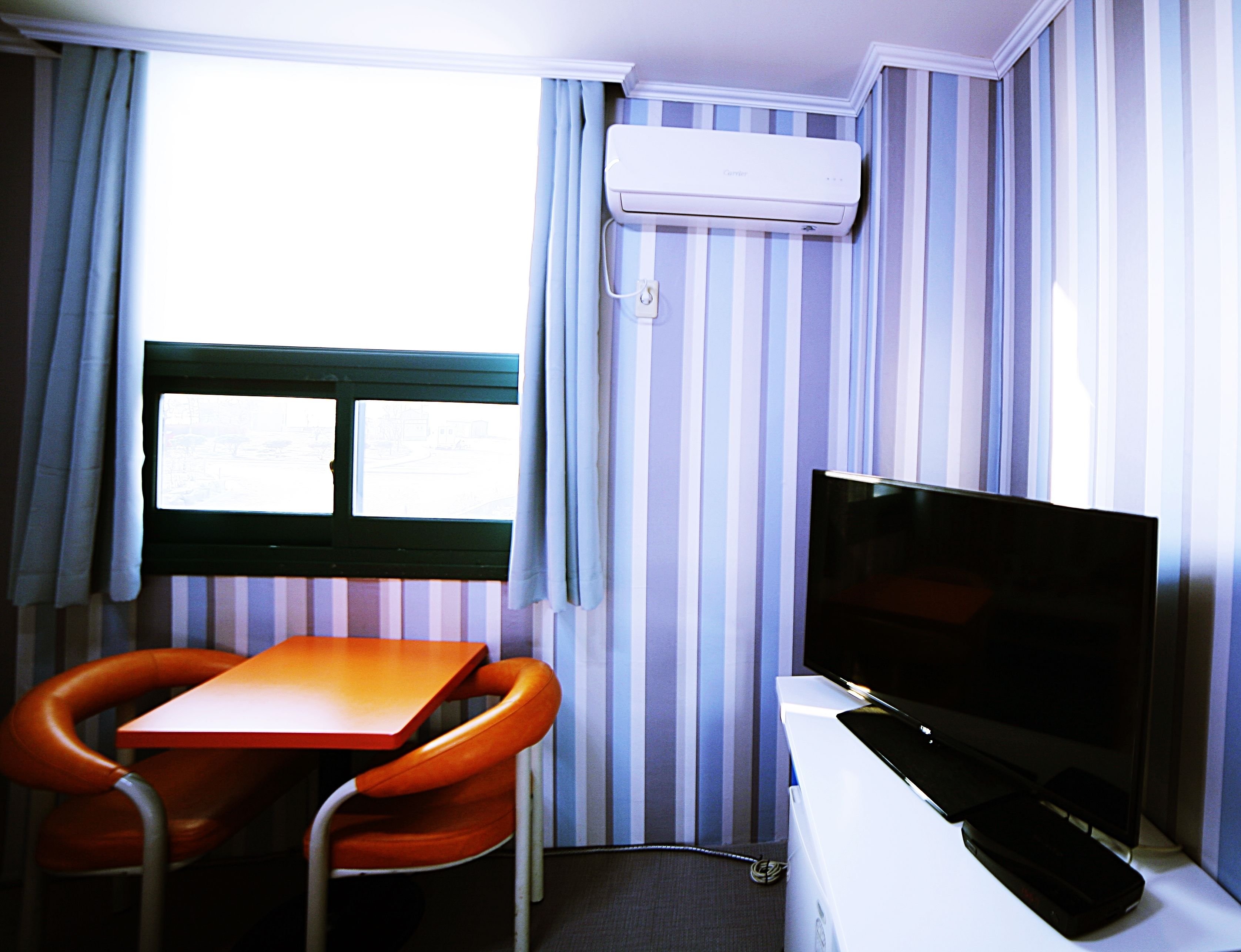 A room with a striped wall, a window with curtains, a television, and a table with a chair.