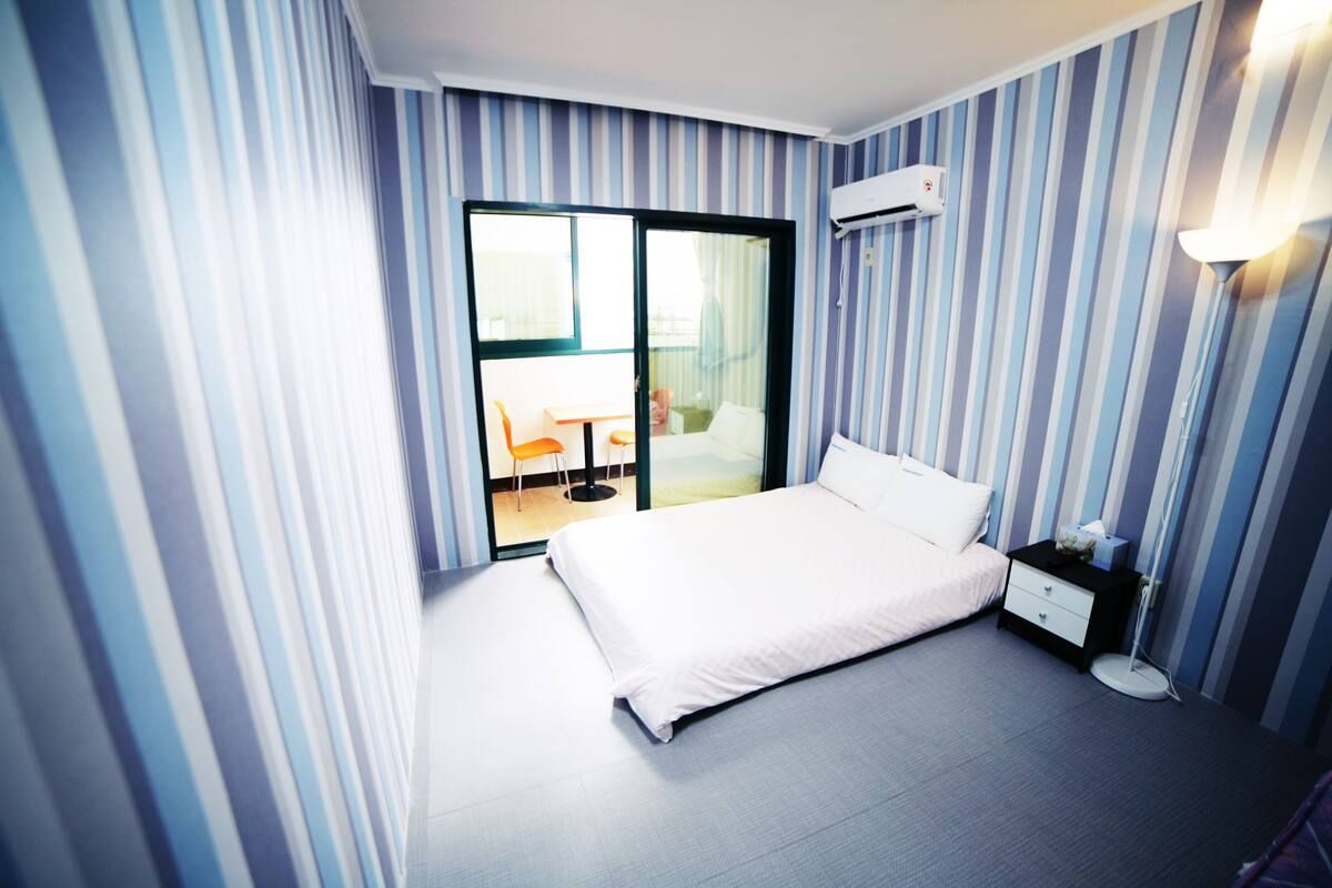 A hotel room with a bed, a nightstand, a lamp, and a view of a balcony with a chair.