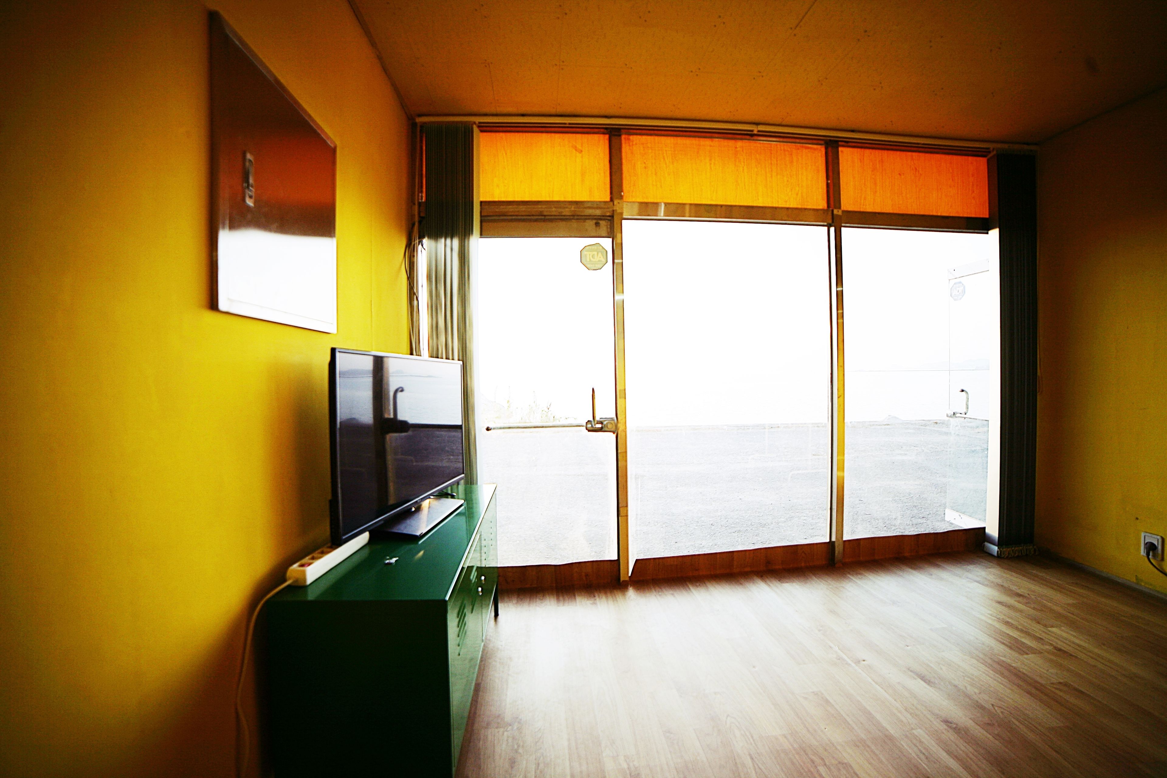 A room with a wooden floor, a green desk with a flat-screen TV, and a sliding glass door leading to a balcony.