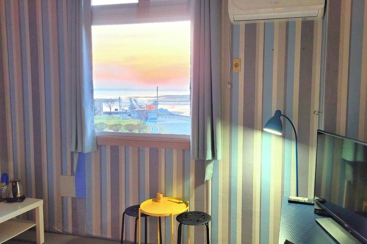A room with striped wallpaper, a window with a view of a landscape, a desk with a lamp, and a television.