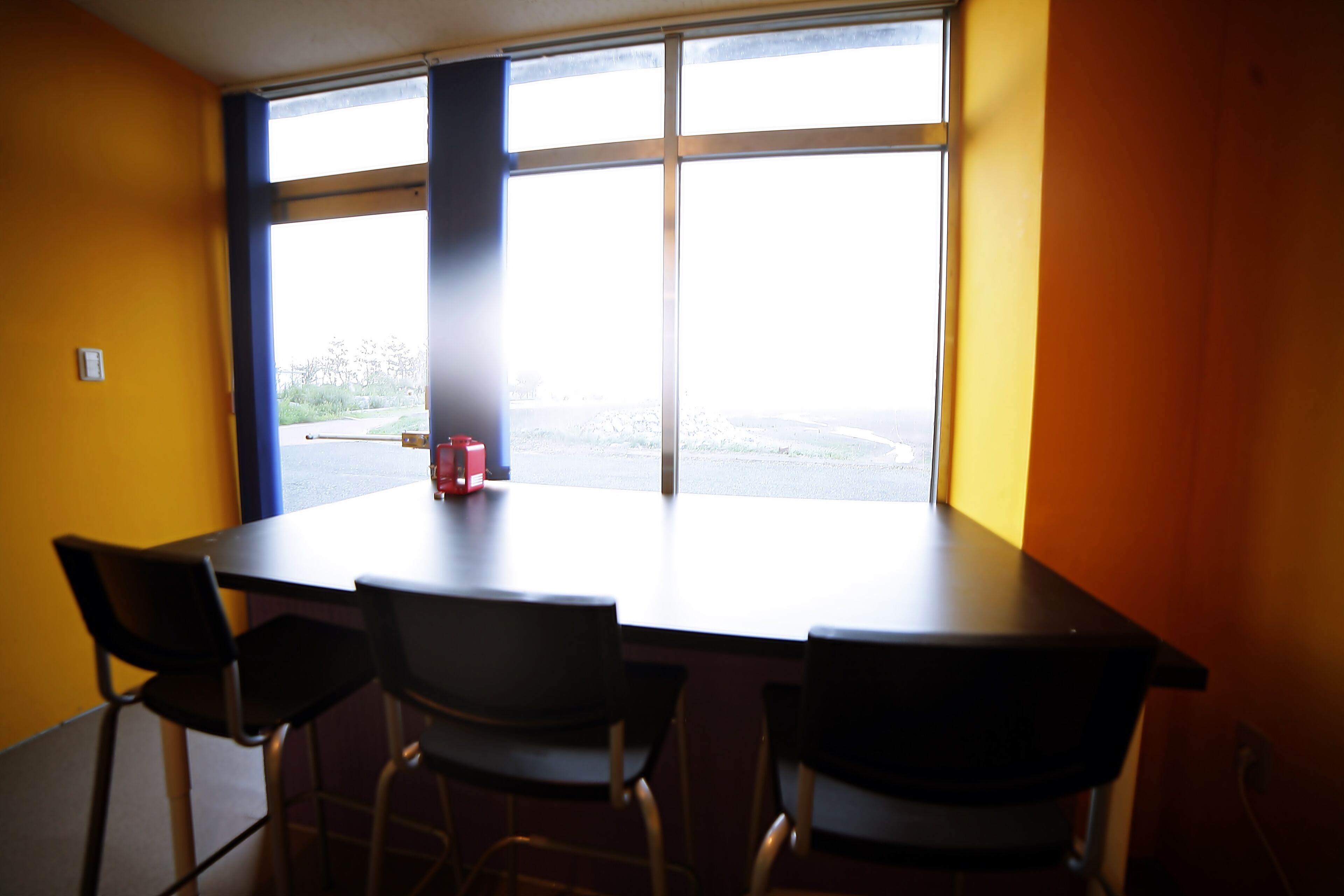 Uma sala de conferências com uma mesa grande e quatro cadeiras, uma janela com vista para um estacionamento e vegetação, e uma parede amarela.