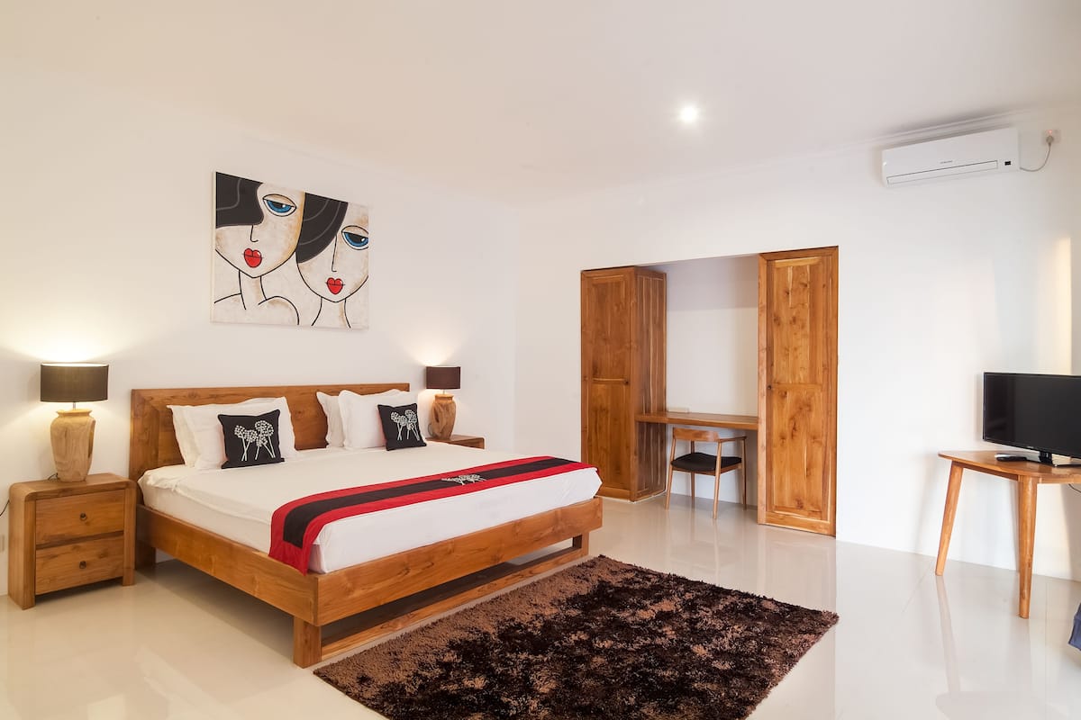 A modern bedroom with a wooden bed, bedside tables, a desk, a chair, a TV, and artwork on the wall.