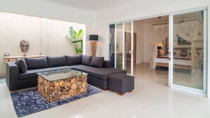 Una sala de estar moderna con un sofá seccional, una mesa de centro de vidrio con base de madera y un amplio ventanal corredera que da a un dormitorio con cama con dosel.