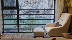 A room with a large window overlooking a rocky cliff and a body of water. There is a white armchair with a cushion and a small ottoman. - Spring Posh Roadsun International Hotel & Resort (Zhangjiajie)