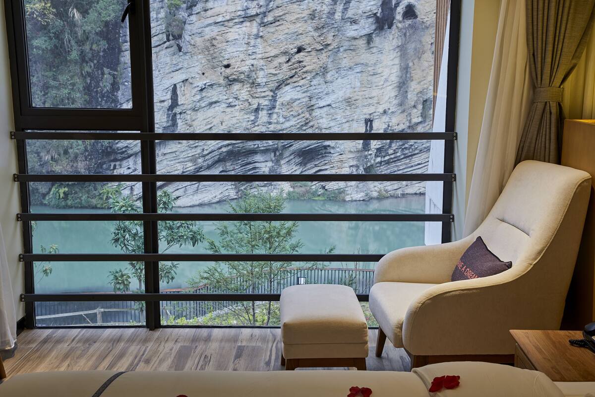 Ein Zimmer mit einem großen Fenster, das auf eine steile Klippe und ein Gewässer blickt. Es gibt einen weißen Sessel mit Kissen und einen kleinen Hocker.