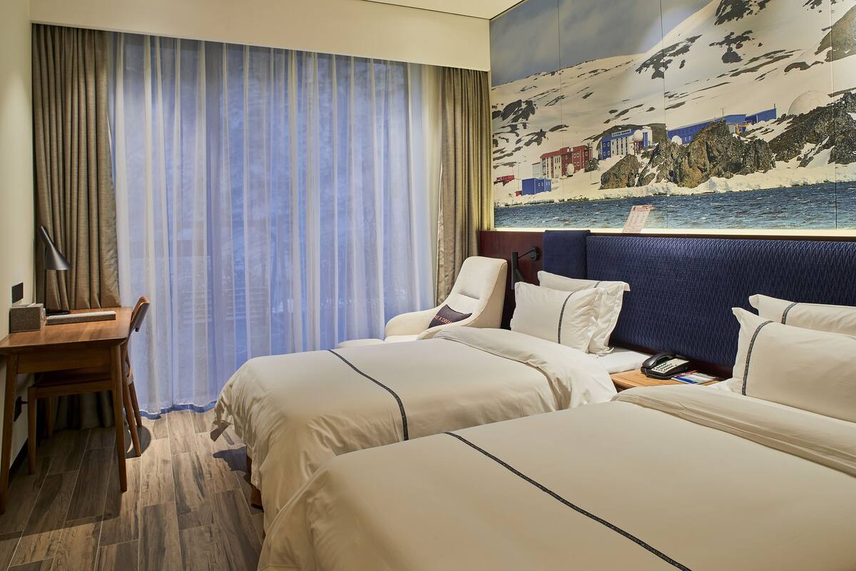 Ein Hotelzimmer mit zwei Betten, einem Schreibtisch, einem Stuhl und einem Gemälde mit einer Küstenansicht.