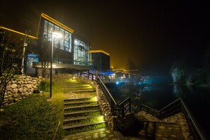 Vista noturna de um edifício moderno com fachada de vidro, degraus de pedra que levam a uma escadaria de madeira, e um corpo de água refletindo luzes.
