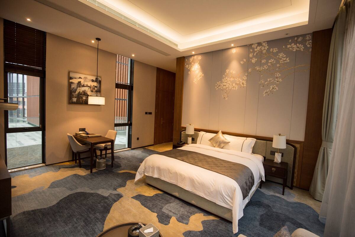 Ein modernes Hotelzimmer mit einem großen Bett, einem Schreibtisch und einem Stuhl. Es gibt ein Fenster mit Jalousien, ein Bild an der Wand und eine dekorative Wandverkleidung.