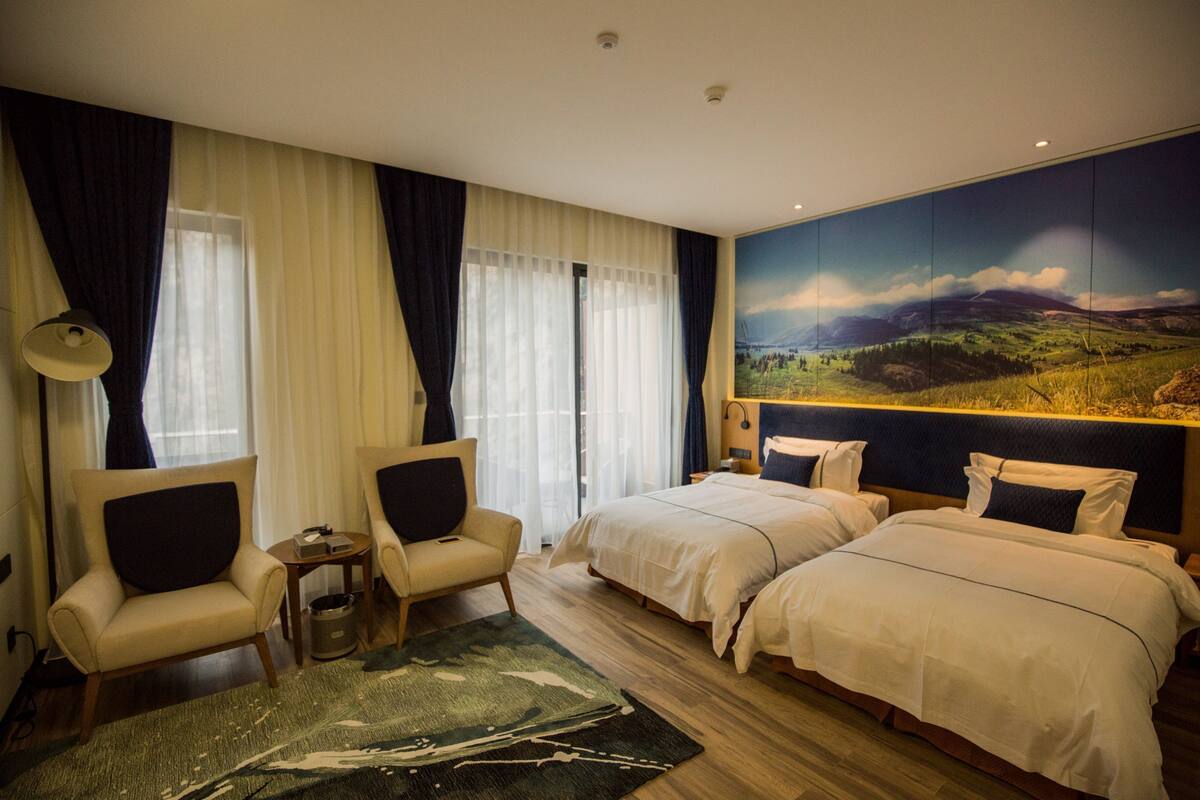 Ein Hotelzimmer mit zwei Betten, einem Sessel, einem kleinen Tisch und einem großen Landschaftsbild an der Wand.