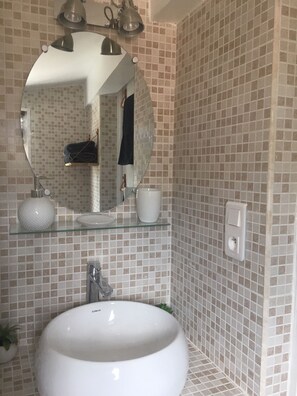 A bathroom with a round white sink, a glass shelf with toiletries, a mirror, and a tiled wall.