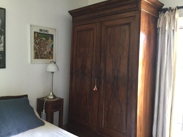 A bedroom with a wooden wardrobe, a bedside table with a lamp, a framed picture on the wall, and a bed with a blue pillow.