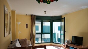 A room with a sofa, a TV on a wooden cabinet, a large window with curtains, and a ceiling light fixture. - Barreiros Apartment - Catedrales Beach 5 pers. (2 rooms) views to the Foz Estuary (Barreiros)