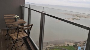Un balcón con mesa y sillas de madera, una planta en maceta y vista a un río y tierra distante.