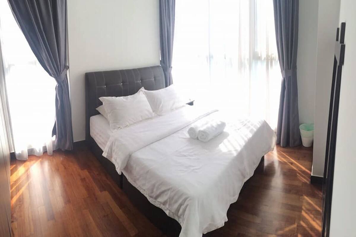 A neatly made bed with white linens and pillows, a wooden floor, and a window with curtains.