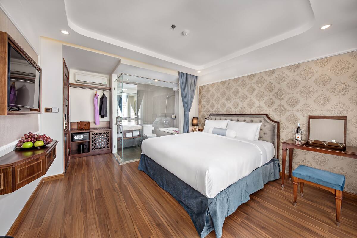 A hotel room with a large bed, wooden flooring, a vanity with a mirror, a small table with a lamp, and a bathroom visible through an open door.