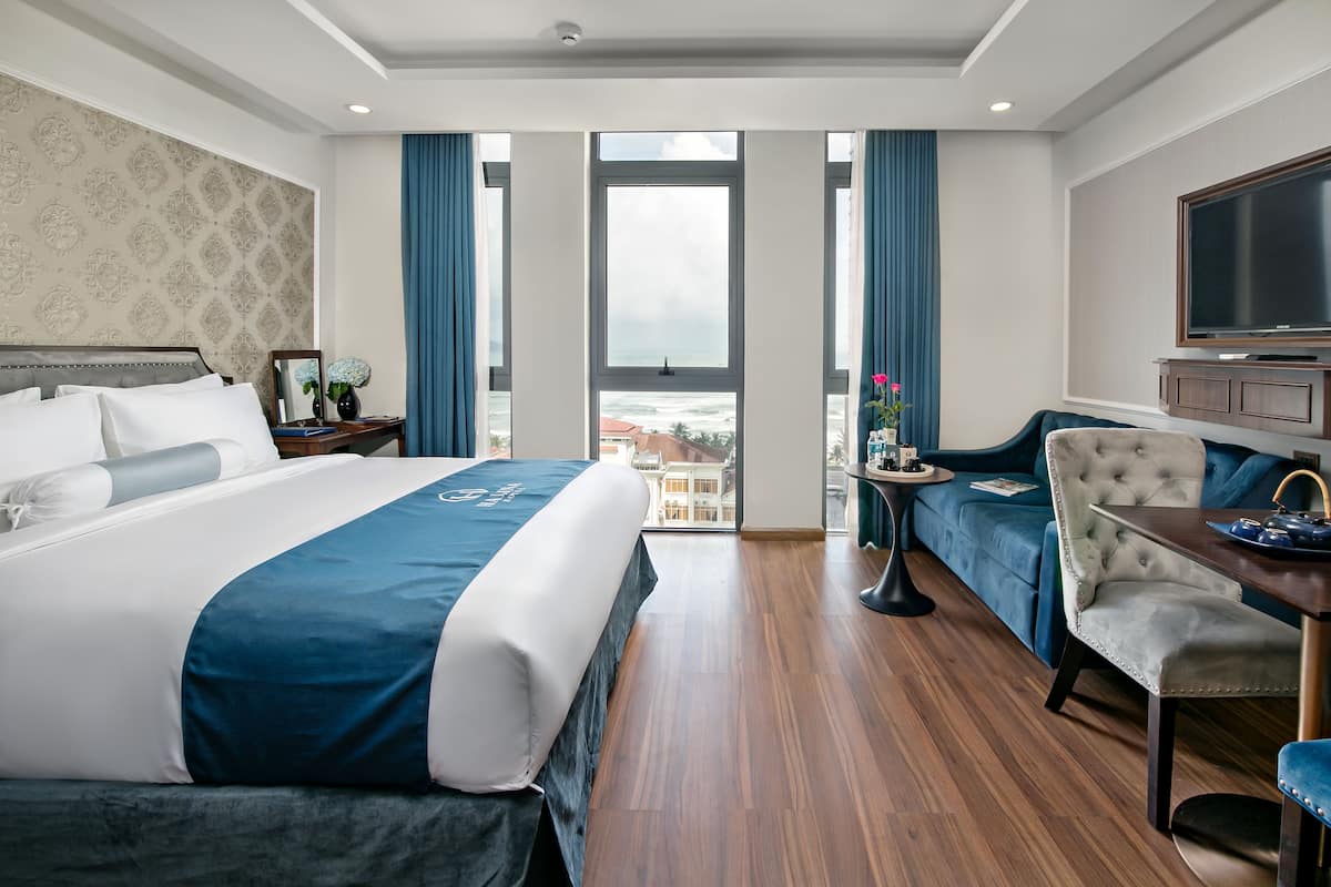 A hotel room with a large bed, a seating area with a sofa and armchairs, a small table, and a TV.